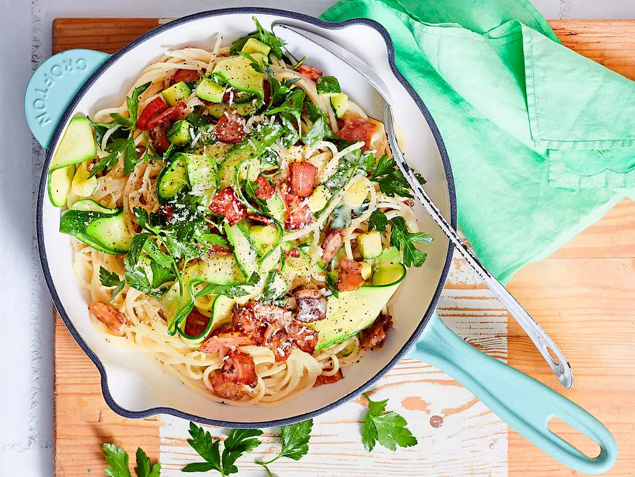 Locker-leichte Zucchini-Carbonara Rezept Spaghetti-Carbonara mit Zucchini und knusprigem Speck in einer hellblauen Pfanne, garniert mit frischen Kräutern und Parmesan