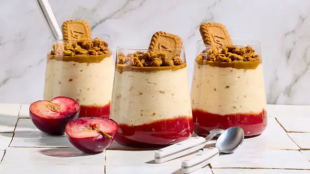 Drei Gläser mit cremigem Lotus-Dessert auf Pflaumenkompott, getoppt mit Keksstückchen und jeweils einem Keks, auf weißen Fliesen serviert, daneben frische, halbierte Pflaumen und zwei silberne Löffel. - Foto: StockFood Studios/Meike Bergmann Drei Gläser mit cremigem Lotus-Dessert auf Pflaumenkompott, getoppt mit Keksstückchen und jeweils einem Keks, auf weißen Fliesen serviert, daneben frische, halbierte Pflaumen und zwei silberne Löffel. - Foto: StockFood Studios/Meike Bergmann
