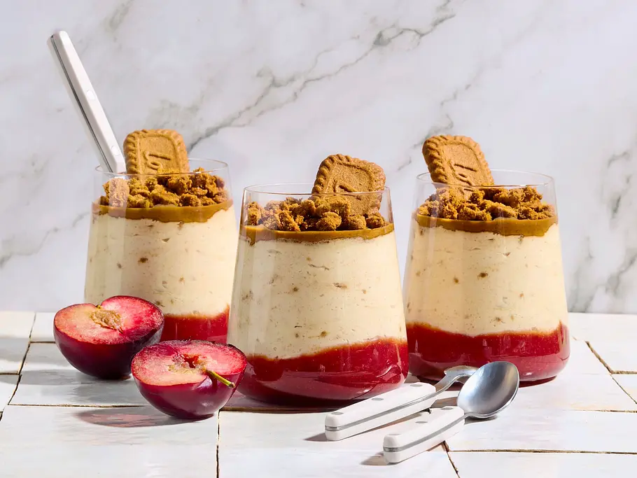 Drei Gläser mit cremigem Lotus-Dessert auf Pflaumenkompott, getoppt mit Keksstückchen und jeweils einem Keks, auf weißen Fliesen serviert, daneben frische, halbierte Pflaumen und zwei silberne Löffel.