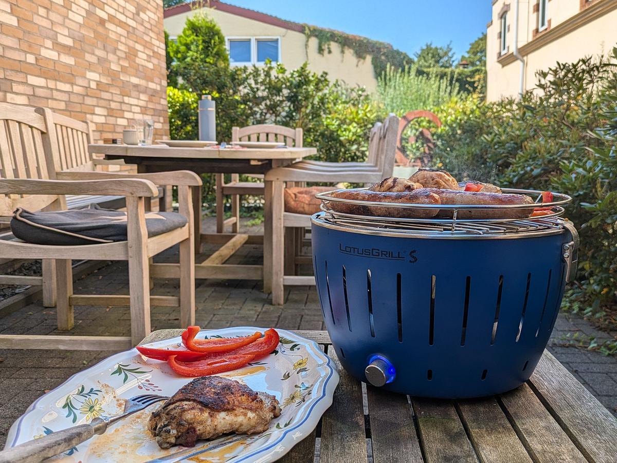Der LotusGrill S steht auf einem Holztisch, darauf Würste und Paprika, im Vordergrund ein Teller mit Grillgut und Besteck.