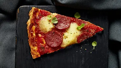 Ein Stück Salami-Pizza mit geschmolzenem Käse und frischer Tomatensauce, belegt mit Salami und ein paar Oreganoblättern, serviert auf einem schwarzen, eckigen Brett. - Foto: LECKER @ Bauer Media Group