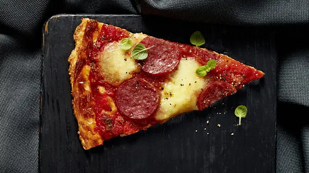 Ein Stück Salami-Pizza mit geschmolzenem Käse und frischer Tomatensauce, belegt mit Salami und ein paar Oreganoblättern, serviert auf einem schwarzen, eckigen Brett. - Foto: LECKER @ Bauer Media Group