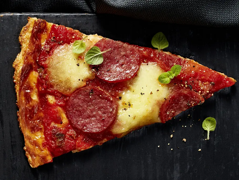 Ein Stück Salami-Pizza mit geschmolzenem Käse und frischer Tomatensauce, belegt mit Salami und ein paar Oreganoblättern, serviert auf einem schwarzen, eckigen Brett.