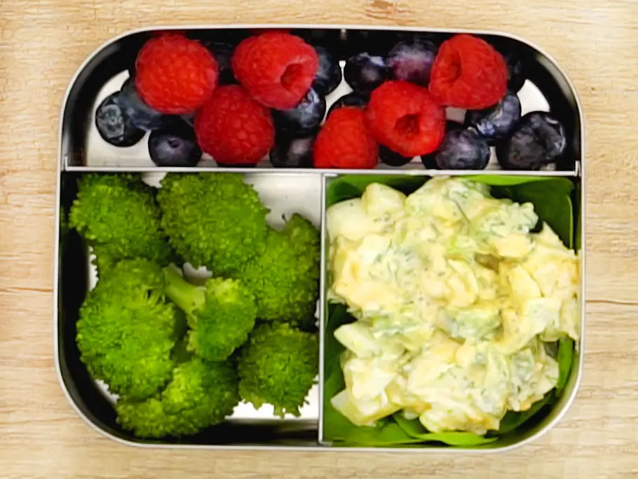 Gekochte Brokkoliröschen, bunte Beerenmischung aus Himbeeren und Heidelbeeren sowie cremiger Eiersalat auf Spinatblättern, sortiert in einer Edelstahl-Lunchbox.