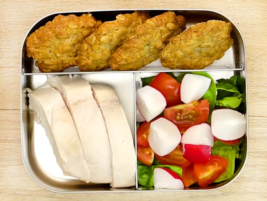 Geschnittenes Hähnchenbrustfilet, knusprige Müslibrötchen sowie ein frischer Salat aus Tomaten, Radieschen und Blättern, appetitlich sortiert in einer Edelstahl-Lunchbox.