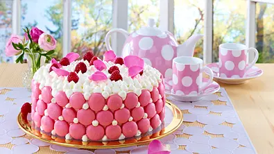 Eine rosa-weiße Torte mit Macarons, Himbeeren und Blütenblättern steht neben gepunktetem Teegeschirr und Blumen auf einem Tisch. - Foto: LECKER @ Bauer Media Group Eine rosa-weiße Torte mit Macarons, Himbeeren und Blütenblättern steht neben gepunktetem Teegeschirr und Blumen auf einem Tisch. - Foto: LECKER @ Bauer Media Group