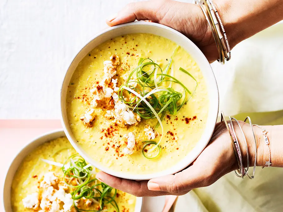 Eine gelbe Maissuppe mit Toppings wird in einer Schüssel von zwei Händen gehalten.