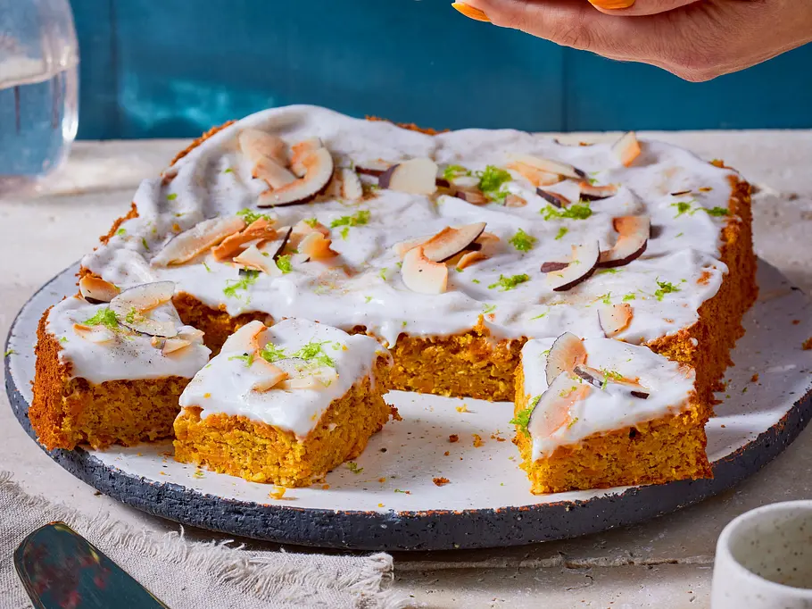 Mandelkuchen mit Limetten-Kokos-Joghurt Hände halten einen türkisfarbenen Teller mit einem Stück Kuchen. Darunter liegt Kuchen mit weißem Guss und Kokoschips.