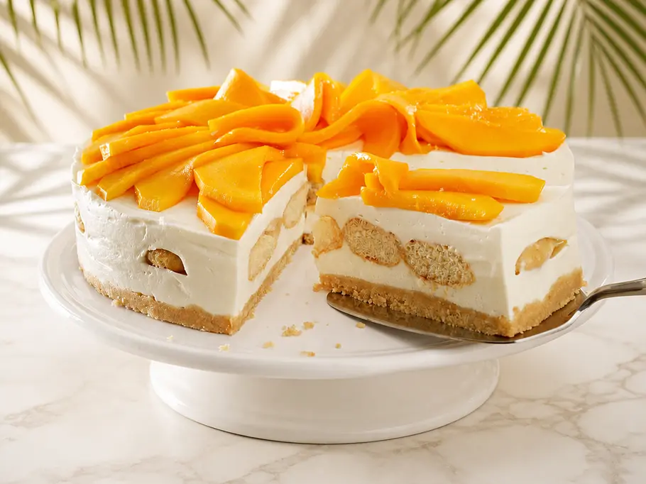 Mango-Tiramisu-Cheesecake Auf einem hellen Marmoruntergrund steht ein cremiger Cheesecake im Tiramisu-Stil mit Löffelbiskuits als Boden und Zwischenschicht und mit Mangoscheiben-Topping auf einem weißen Tortenständer.