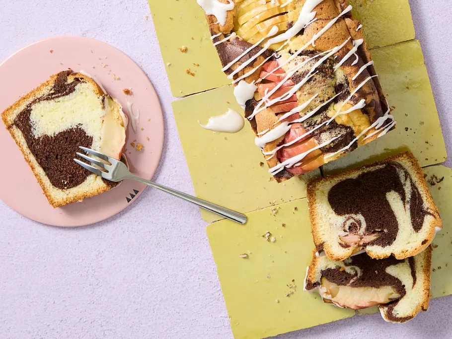 Marmor-Apfelkuchen Rezept Saftiger Marmor-Apfelkuchen als Kastenkuchen mit dunkler und heller Teigschicht, Apfelstücken und Zuckerguss, in Scheiben geschnitten auf einem gelben Untergrund.