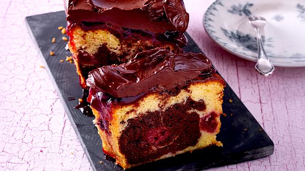 Angeschnittener Marmor-Kirsch-Kuchen aus der Kastenform mit üppiger Schoko-Ganache mit Kuchenteller im Hintergrund. - Foto: House of Food / Bauer Food Experts KG