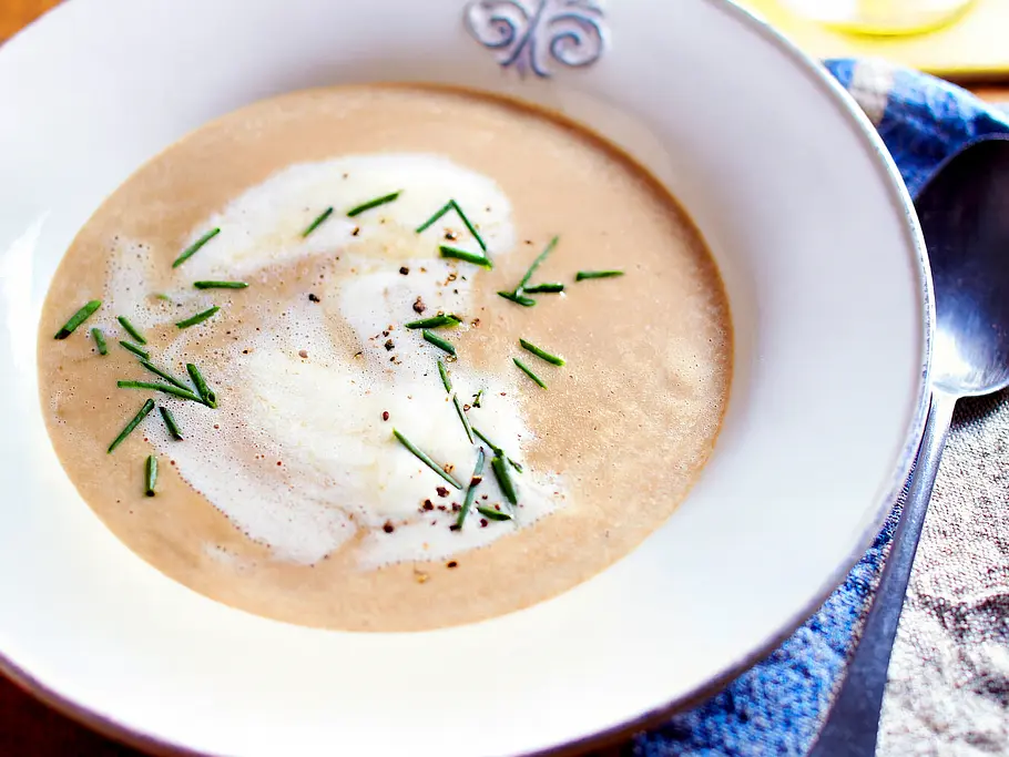 Maronensuppe Rezept Ein weißer Suppenteller gefüllt mit cremiger Maronensuppe, garniert mit aufgeschäumter Sahne, grobem Pfeffer sowie frischem Schnittlauch