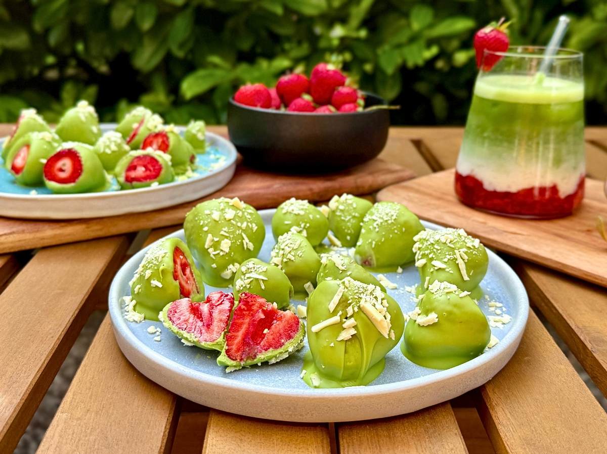 Erdbeeren umhüllt von Matcha-Schokolade auf Tellern mit frischen Erdbeeren und einem Iced Strawberry Latte im Hintergrund
