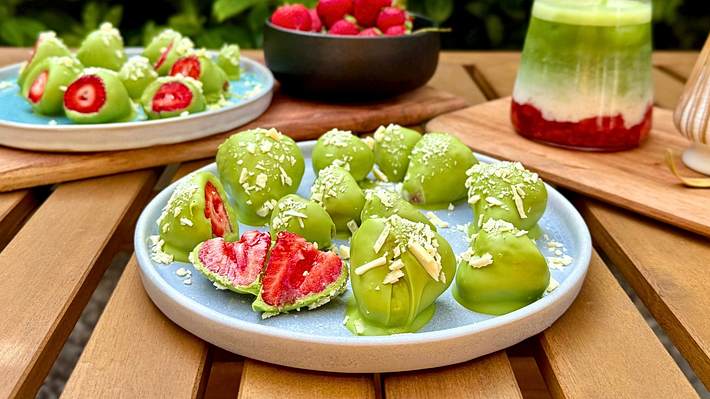Erdbeeren umhüllt von Matcha-Schokolade auf Tellern mit frischen Erdbeeren und einem Iced Strawberry Latte im Hintergrund - Foto: Franziska Hoppe / LECKER.de