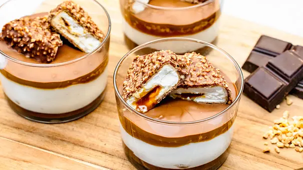 Drei Gläser mit geschichtetem Cremedessert und Schokoriegelstücken stehen neben Schokolade und gehackten Nüssen auf einem Holzbrett. - Foto: ShowHeroes für LECKER @ Bauer Media Group