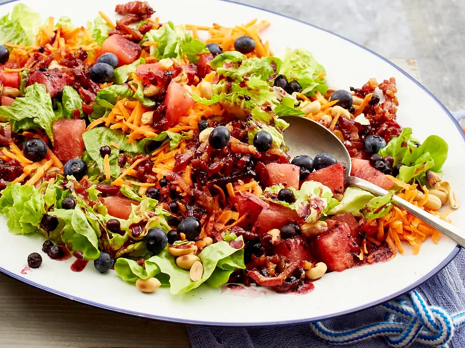  Bunte Blattsalate, Möhren, Wassermelone, Blaubeeren, Erdnüsse und knuspriger Speck liegen angerichtet auf einer weißen Platte.
