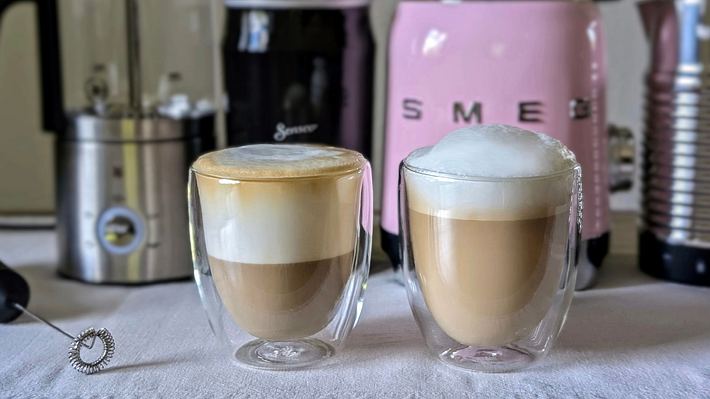 Mehrere Milchaufschäumer stehen nebeneinander auf einem Esstisch, davor zwei Gläser Cappuccino – mit unterschiedlich dicker Schaumhaube. - Foto: LECKER.de / Constanze Brinkschulte