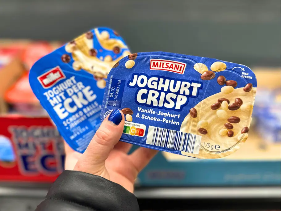 Eine Hand hält eine Packung Milsani Joghurt Crisp und einen Joghurt mit der Ecke von Müller