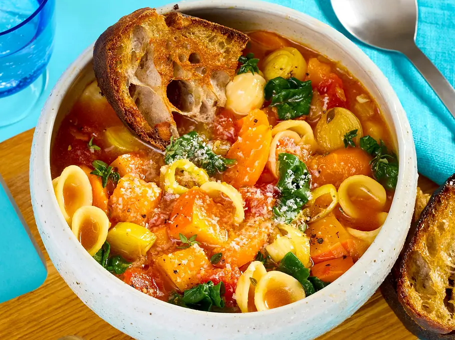Bunte Minestrone mit Hokkaido-Kürbis, Spinat und kleinen Nudeln in einer weißen Schale, serviert auf einem Holzbrett mit knusprigem Brot, Löffel und blauer Serviette.