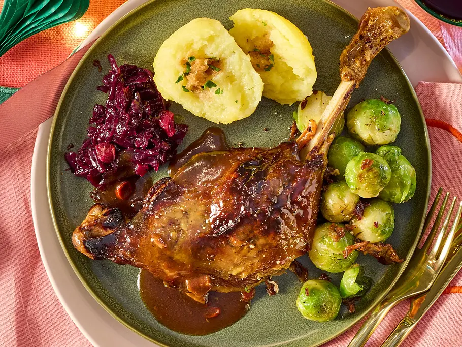 Saftige Gänsekeule mit glänzender Miso-Glasur und Bratensauce auf einem grünen Teller, dazu zwei Kartoffelklöße, Rotkohl und gebratener Rosenkohl auf rosa Stoffserviette.