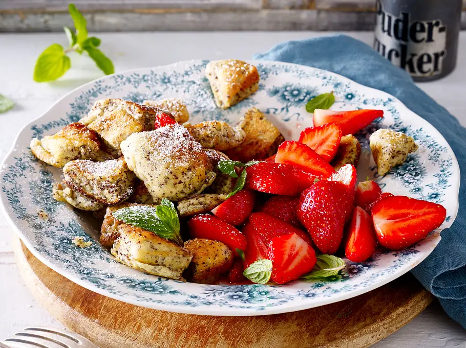 Mohnschmarrn aus dem Airfryer mit frischen Erdbeeren Rezept Luftiger Mohnschmarrn mit Puderzucker bestäubt, dazu frische, geviertelte Erdbeeren auf einem gemusterten Porzellanteller.