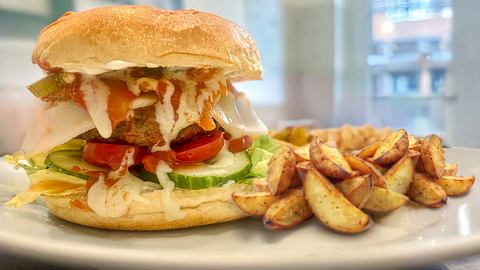 Ein Brioche-Bun-Burger üppig belegt mit Rindfleisch-Patty, Kopfsalat, Gurke, Tomate, Ziegenkäse, Jalapenos und Kimchi, goldbraune Kartoffelecken als Beilage - Foto: Elena S.