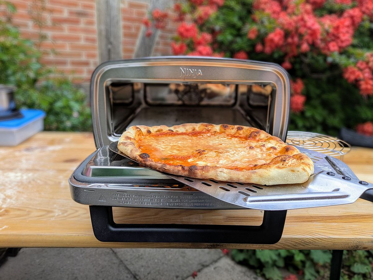 Eine goldbraun gebackene Pizza Margherita wird mit einem Schieber aus dem Ninja Artisan Outdoor-Elektro-Pizzaofen geholt.