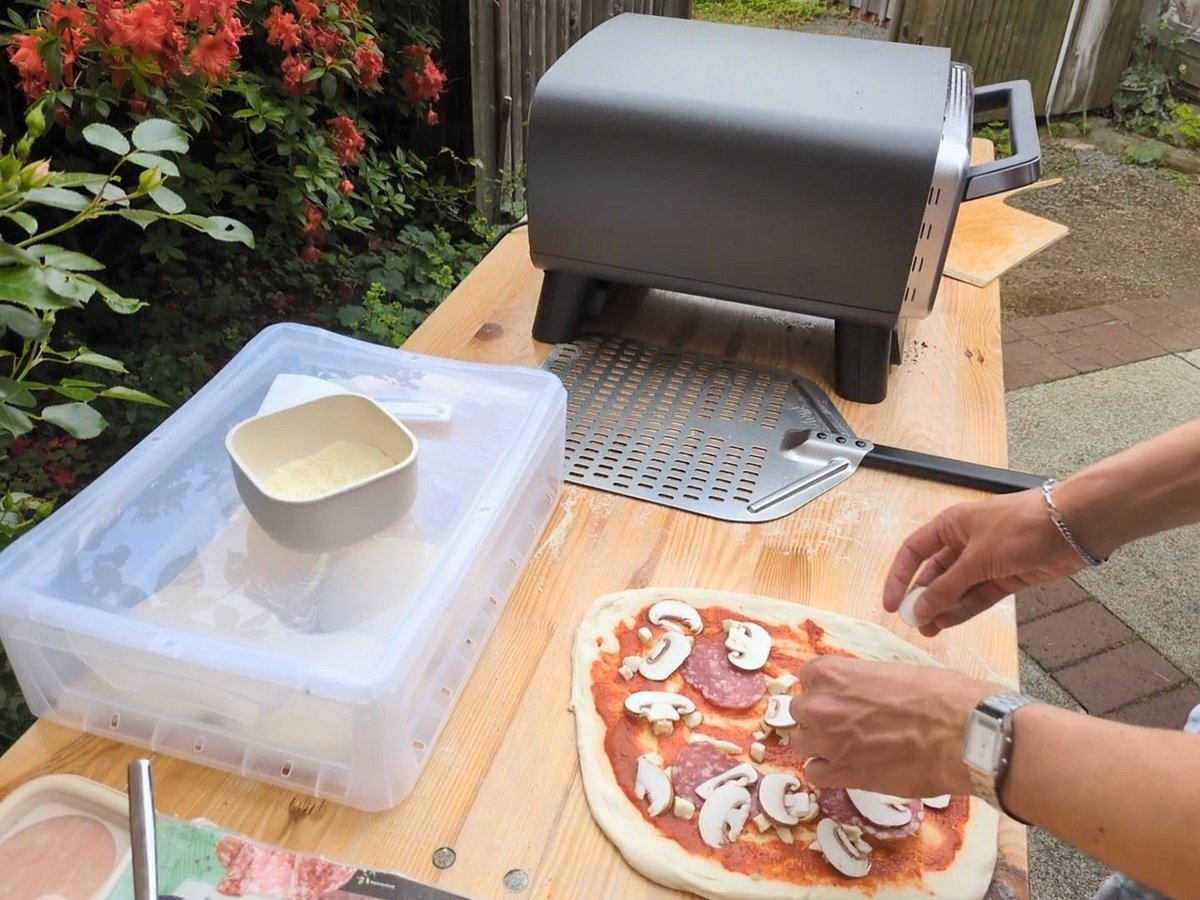 ninja-artisan-pizzaofen-belegen Eine Person belegt eine rohe Pizza im Freien neben dem Ninja Artisan Outdoor-Elektro-Pizzaofen auf einem Holztisch.