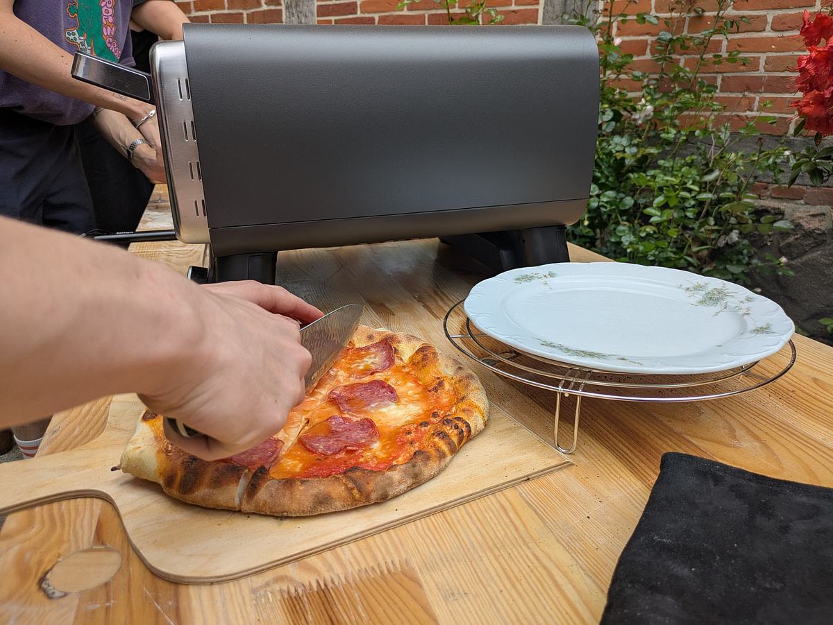 ninja-artisan-pizzaofen-salami-pizza Eine Pizza Salami wird neben dem Ninja Artisan Pizzaofen auf einem Holzbrett in Stücke geschnitten.