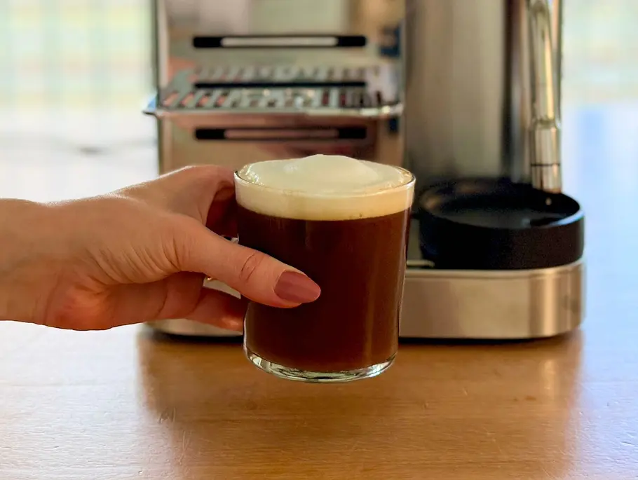 Ninja AutoBarista Pro Cold Brew Ein Cold Brew mit weißem Milchschaum in einem Glas gehalten von einer Hand vor der Ninja AutoBarista Pro
