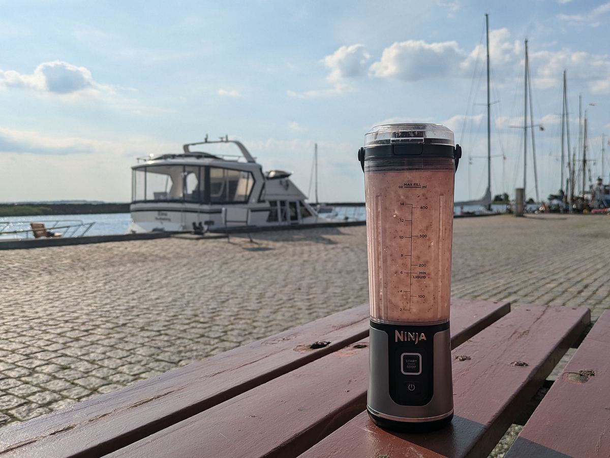 Ninja Blast Smoothie-Maker Der Ninja Smoothie-Maker steht mit blassrotem Smoothie auf einer Holzbank am Hafen, im Hintergrund liegen Boote im Wasser