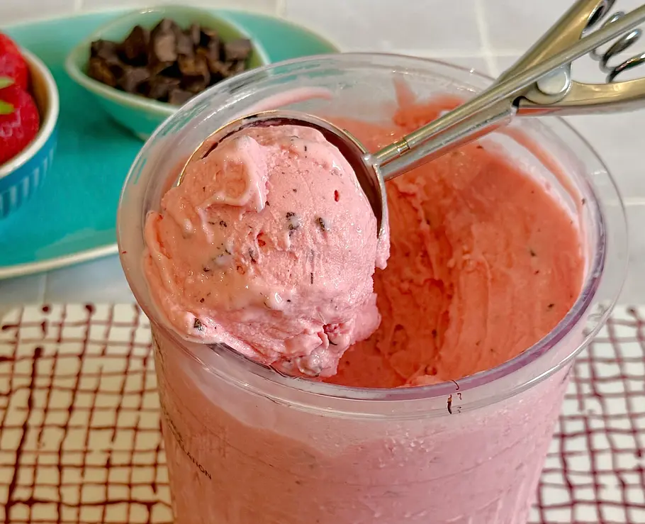 Ninja Creami: Frozen Joghurt im Yogurette-Style Rezept Erdbeer-Frozen-Joghurt mit Schokoladenstücken in einem Zubereitungsbecher der Ninja Creami. Frische Erdbeeren und Schokolade im Hintergrund
