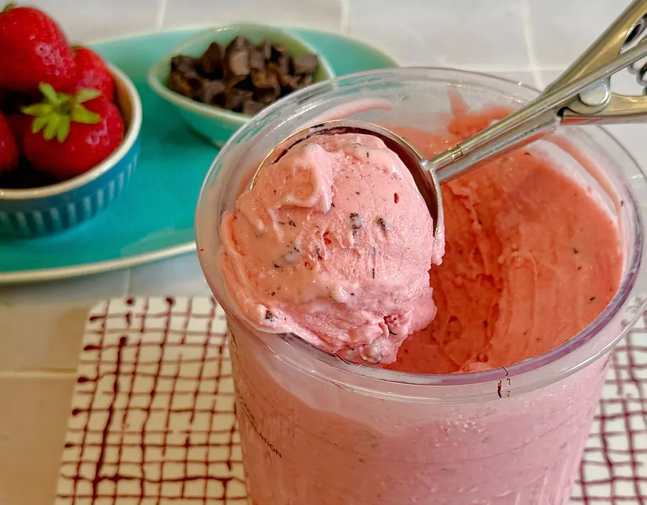 Ninja Creami: Frozen Joghurt im Yogurette-Style Rezept Erdbeer-Frozen-Joghurt mit Schokoladenstücken in einem Zubereitungsbecher der Ninja Creami. Frische Erdbeeren und Schokolade im Hintergrund