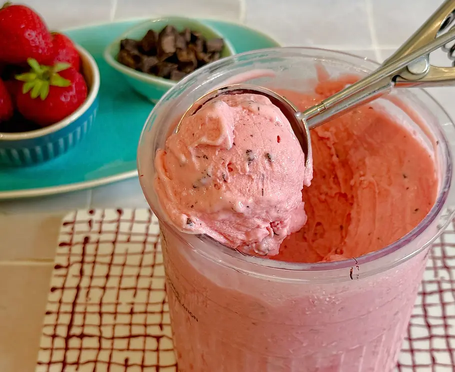 Erdbeer-Frozen-Joghurt mit Schokoladenstücken in einem Zubereitungsbecher der Ninja Creami. Frische Erdbeeren und Schokolade im Hintergrund