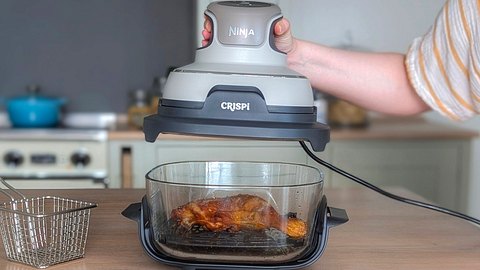 Zu sehen ist ein Arm, dessen Hand den PowerPod des Ninja Crispi Airfryers hochhebt. Darunter steht der Glasbehälter, in dem ein gebratener Hähnchenschenkel liegt. Im Vordergrund stehen Pommes auf einem Teller. - Foto: Constanze Brinkschulte / LECKER.de