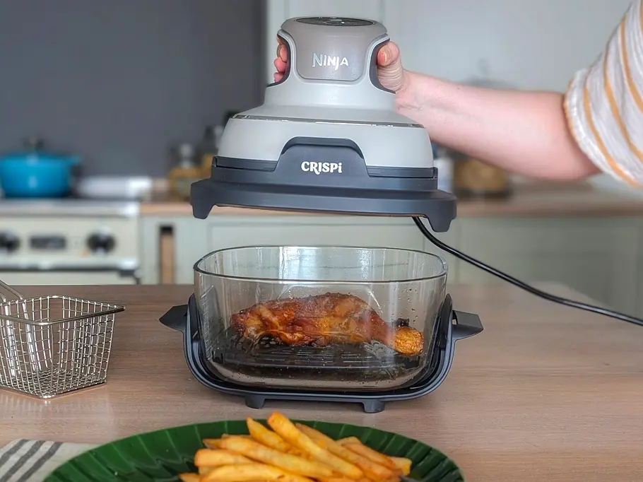 Zu sehen ist ein Arm, dessen Hand den PowerPod des Ninja Crispi Airfryers hochhebt. Darunter steht der Glasbehälter, in dem ein gebratener Hähnchenschenkel liegt. Im Vordergrund stehen Pommes auf einem Teller.