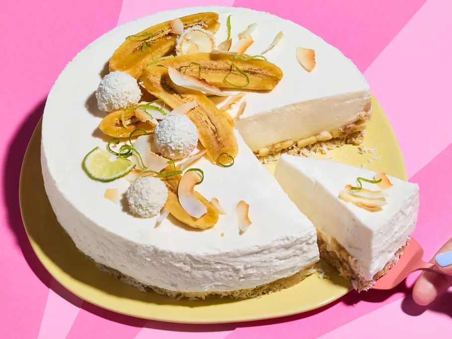 No-Bake-Kokos-Bananen-Cheesecake Eine Hand hebt ein Stück aus einer weißen Torte mit Bananen, Kokos und Limette auf gelbem Teller vor pinkem Hintergrund.