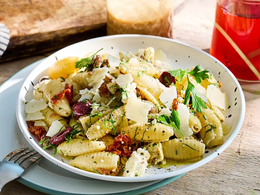  Bunter Nudelsalat mit Feta, Softtomaten, Kalamata-Oliven und frischen Kräutern auf einem weißen Teller, serviert auf rustikalem Holztisch.