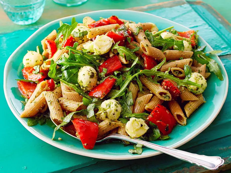 Ein türkiser Teller mit buntem Nudelsalat aus Penne, Rucola, eingelegter Paprika und Mozzarellawürfeln, die mit einem kräuterigen Pesto-Dressing angemacht sind.