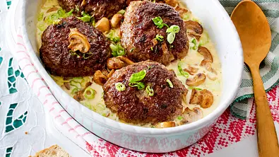 Ofenfrikadellen in Champignon-Lauchzwiebelrahm in einer weißen Auflaufform, daneben ein Holzlöffel. - Foto: LECKER @ Bauer Media Group Ofenfrikadellen in Champignon-Lauchzwiebelrahm in einer weißen Auflaufform, daneben ein Holzlöffel. - Foto: LECKER @ Bauer Media Group