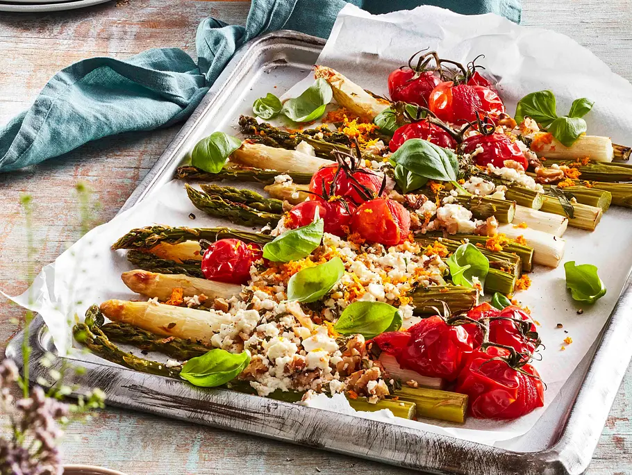 Ofenspargel mit gerösteten Kirschtomaten und zerbröseltem Feta auf einem Backblech, garniert mit frischem Basilikum.