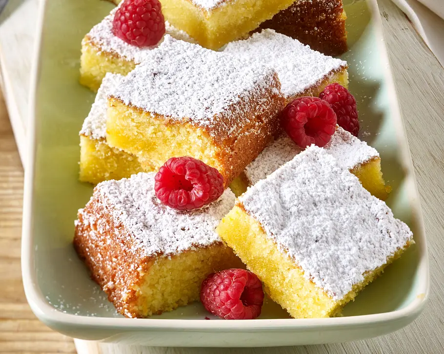 Olivenölkuchen mit Orange Rezept Mehrere Stücke Olivenölkuchen mit Puderzucker bestäubt und frischen Himbeeren auf einer hellen Platte angerichtet.