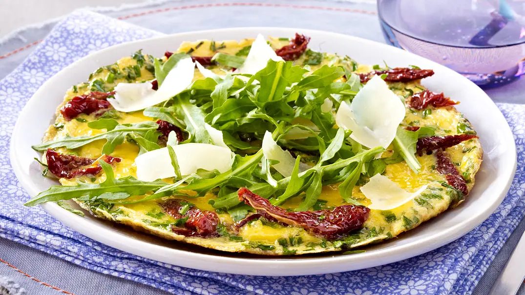 Serviertes Kräuteromelett mit getrockneten Tomaten, Rucola und Parmesanspänen auf einem weißen Teller, darunter ein gemustertes Stofftuch. - Foto: LECKER @ Bauer Media Group Serviertes Kräuteromelett mit getrockneten Tomaten, Rucola und Parmesanspänen auf einem weißen Teller, darunter ein gemustertes Stofftuch. - Foto: LECKER @ Bauer Media Group