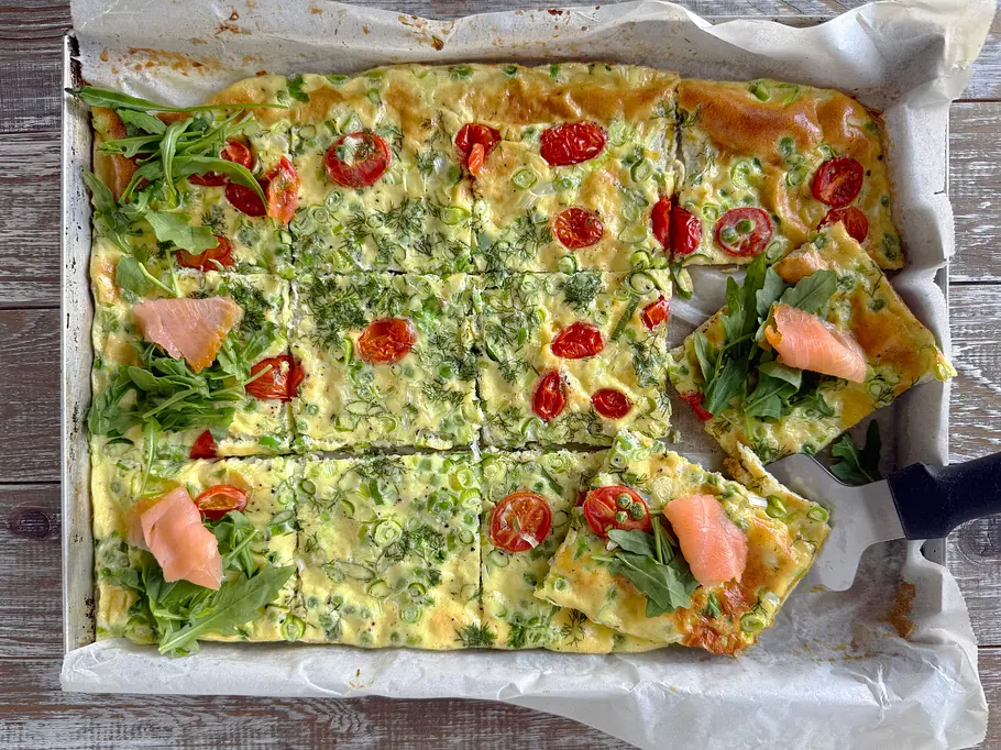 Omelett vom Blech Rezept Auf einem Backblech liegt ein in Stücke geschnittenes Omelett mit Tomaten und Frühlingszwiebeln, teils mit Rucola und Lachs belegt.