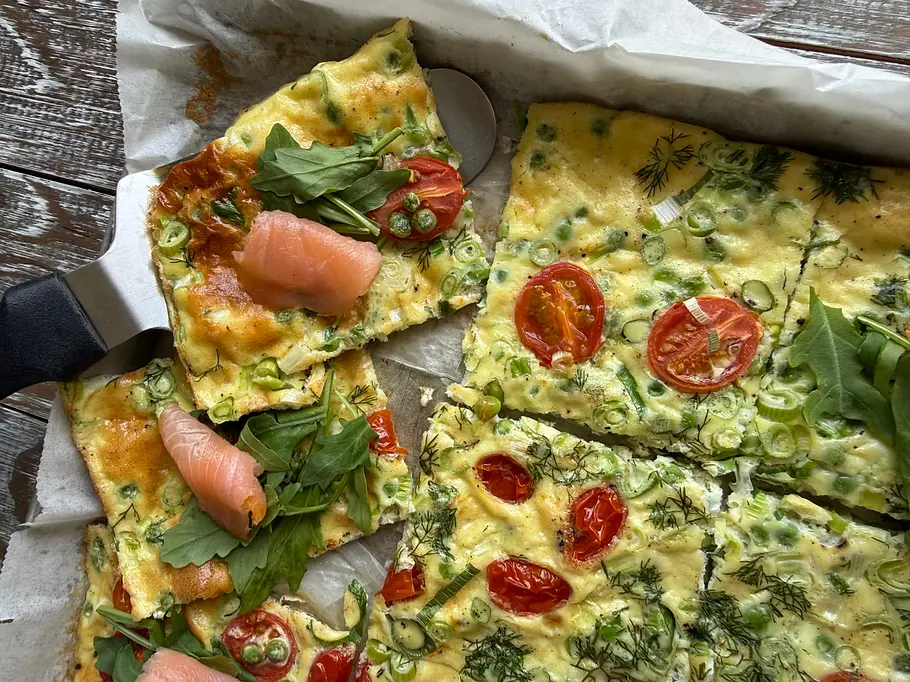 Geniales Brunch-Omelett vom Blech – perfekt für viele Ein in Stücke geschnittenes Ofenomelett mit Tomaten, Frühlingszwiebeln und Kräutern liegt mit Lachs und Rucola auf Backpapier.
