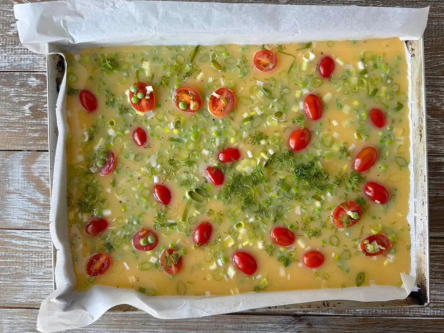 Omelett vom Blech Rezept Auf einem mit Backpapier ausgelegten Blech liegt die flüssige Ei-Masse mit Kirschtomaten, Frühlingszwiebeln und Kräutern.