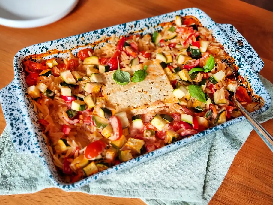 One Pot Ofen-Kritharaki Rezept Auflaufform mit Kritharaki, Zucchini, Paprika und Tomaten. In der Mitte Feta-Käse und garniert mit Basilikum.