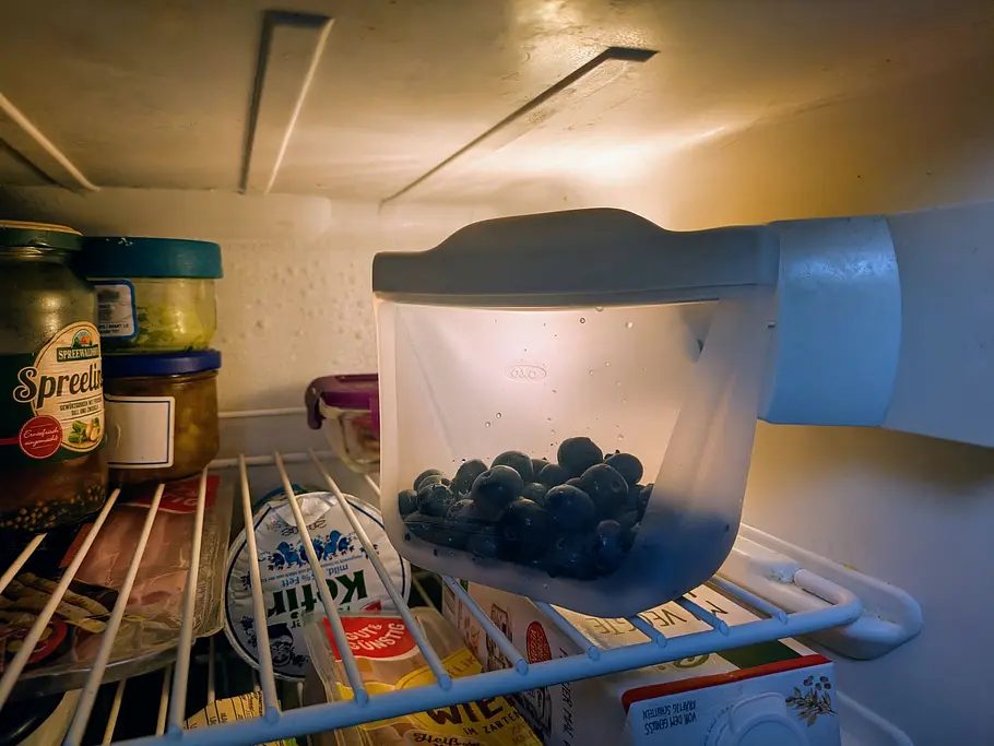 Ein wiederverwendbarer OXO Silikonbeutel mit Blaubeeren steht im beleuchteten Kühlschrank neben weiteren Vorratsbehältern.