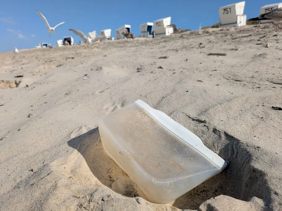 Ein transparenter Silikonbeutel liegt halb im Sand am Strand, im Hintergrund sind Möwen und mehrere weiße Strandkörbe zu sehen.