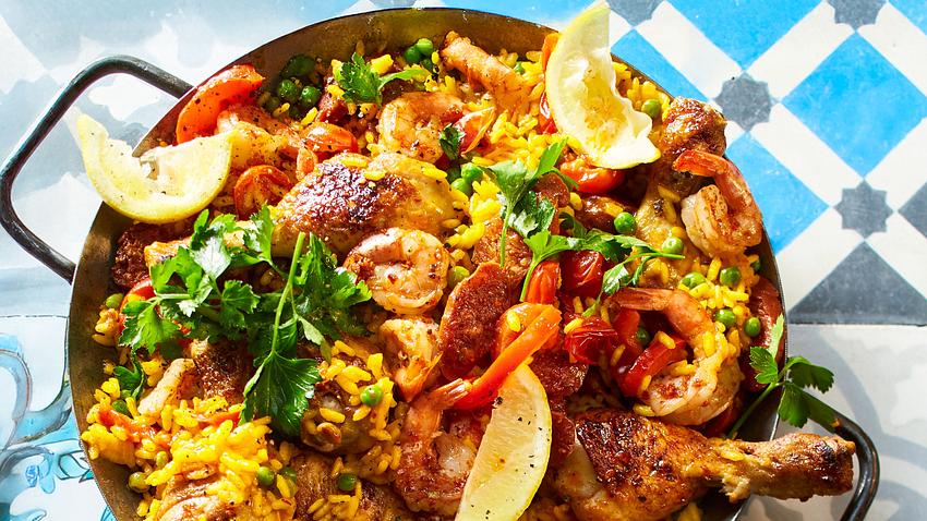 Spanische Paella mit gelbem Reis, Hähnchenkeulen, Garnelen, Erbsen, Paprika und Zitronenspalten, serviert in einer großen Pfanne mit Petersilie garniert. - Foto: House of Food / Bauer Food Experts KG